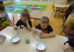 Dwoje dzieci miesza łyżeczkami w słoiku, obserwują zjawisko rozpuszczania soli w wodzie.